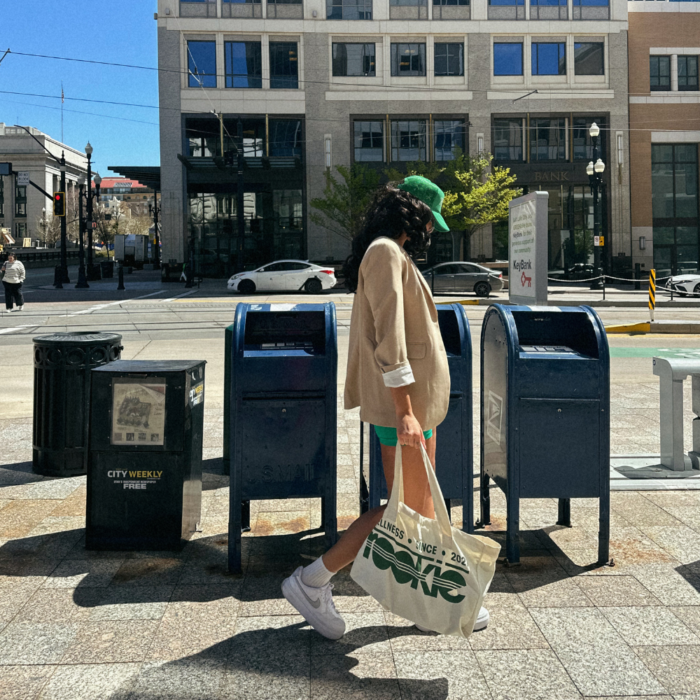 Cream canvas tote bag with "Rookie Est. 2021" text in green, worn by person in urban setting.
