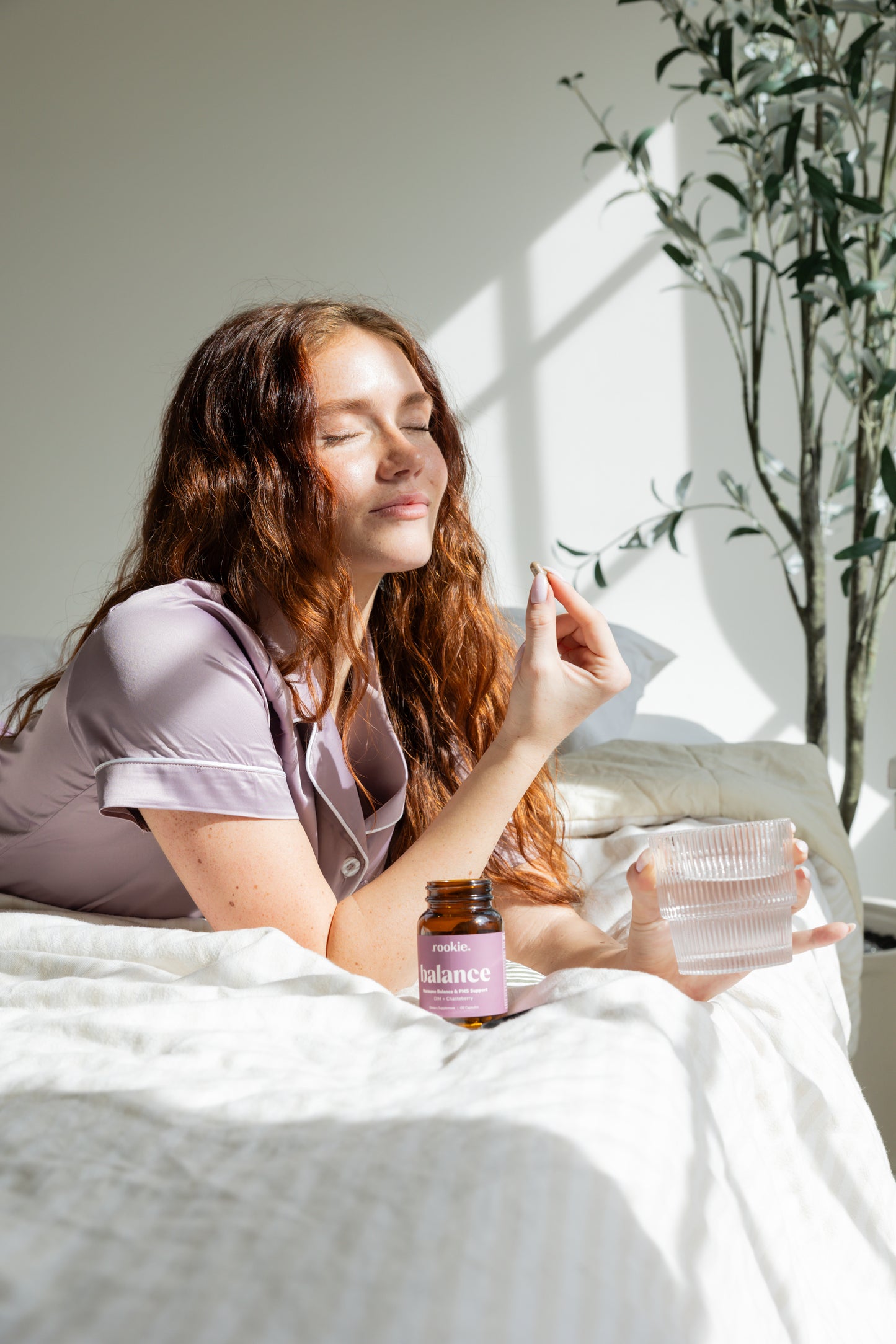 Woman holding a small amber bottle labeled "balance" while sitting on a bed near a potted plant.