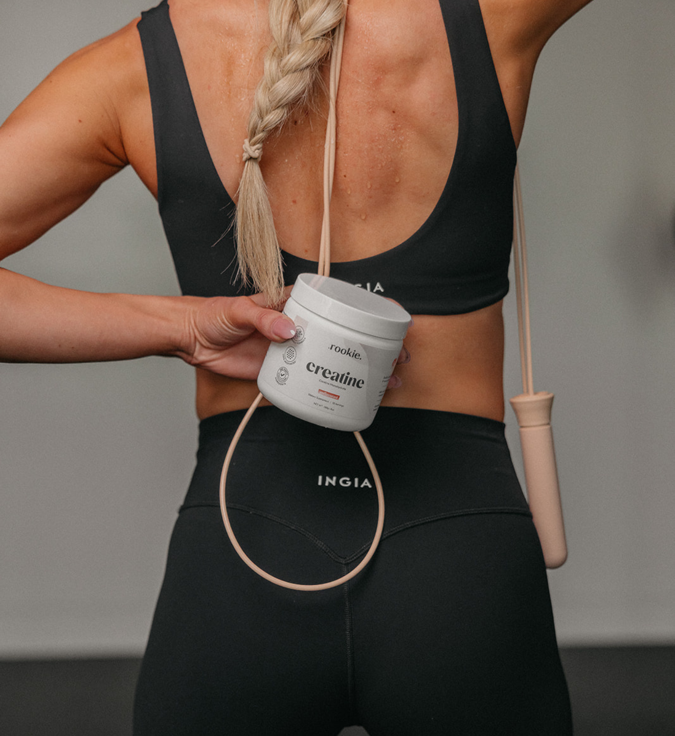 Person holding a container of creatine with a skipping rope, wearing a black sports bra and leggings.