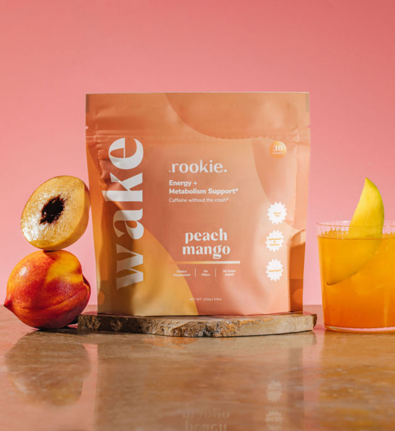 Wake .rookie. peach mango energy supplement pouch displayed with fresh peach, sliced fruit, and orange beverage on pink backg