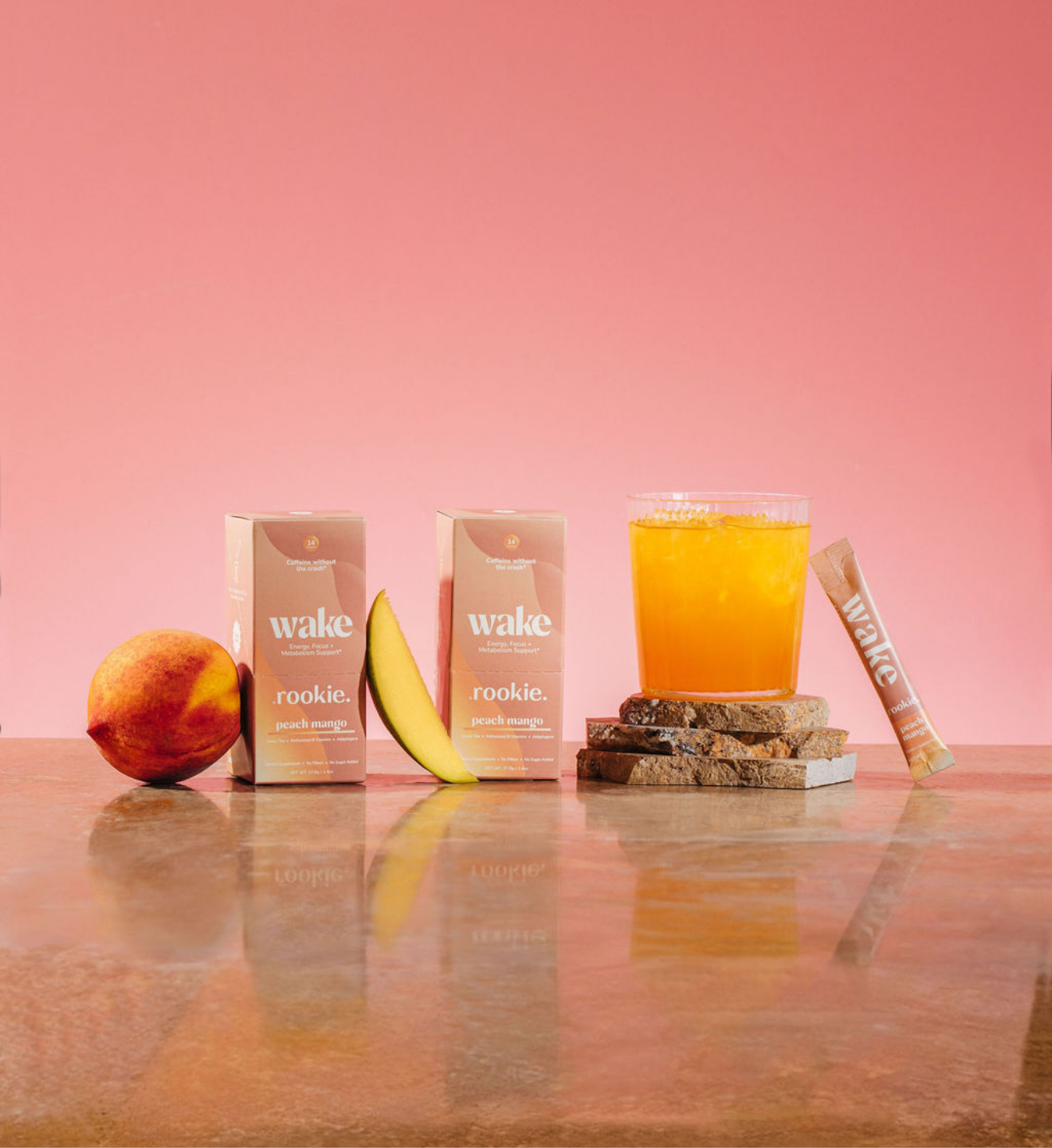Wake rookie peach mango juice boxes, fresh peach, banana slice, glass of juice, and stick displayed against pink background.