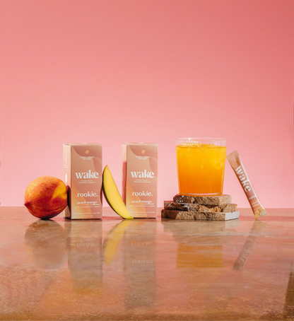 Wake rookie peach mango juice boxes, fresh peach, banana slice, glass of juice, and stick displayed against pink background.